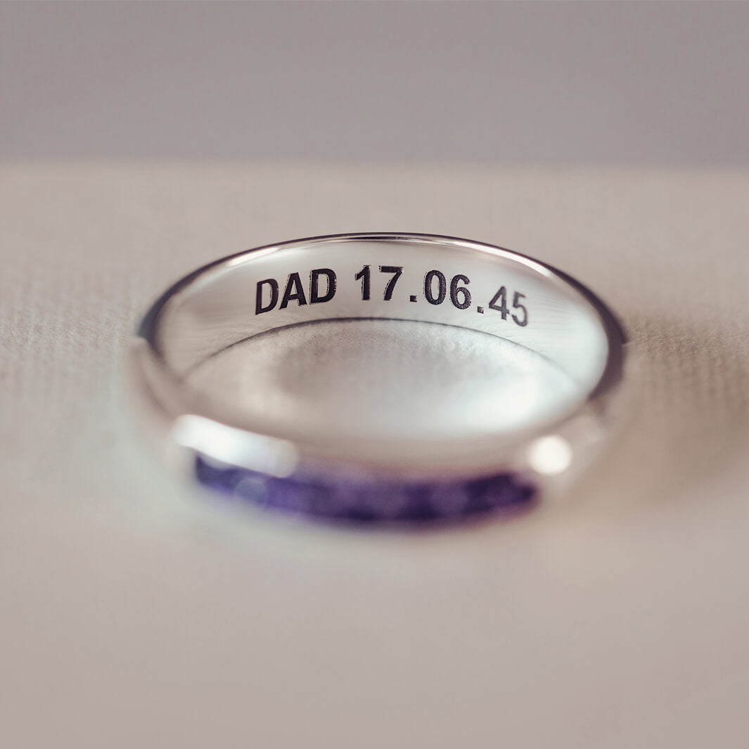 Sterling silver womens ring band with a central section filled with  memorial ashes and purple crushed crystals, on a blurred pale background. The ring has the message 'DAD 17.06.45' engraved into the inside of the ring band in black