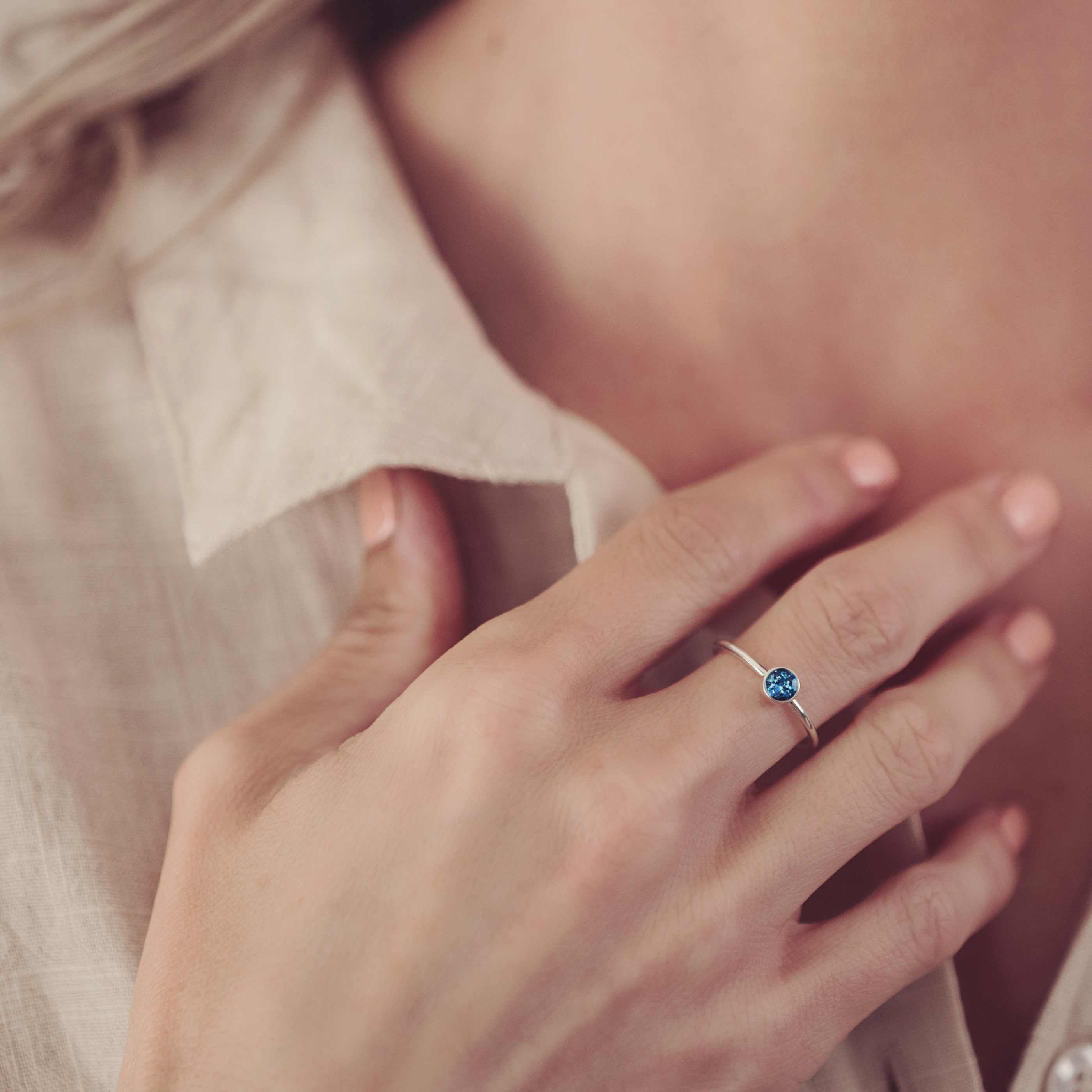 The image shows a close-up of a person’s hand resting gently near their collarbone. They are wearing a delicate ring with a simple band and a small, round blue stone set in a bezel. The stone contains cremation ashes and resin, giving the ring a sentimental and meaningful touch. The person is dressed in a light-coloured shirt, and the soft focus and lighting highlight the ring, drawing attention to its elegance and personal significance. The overall atmosphere is calm and intimate, suggesting the ring