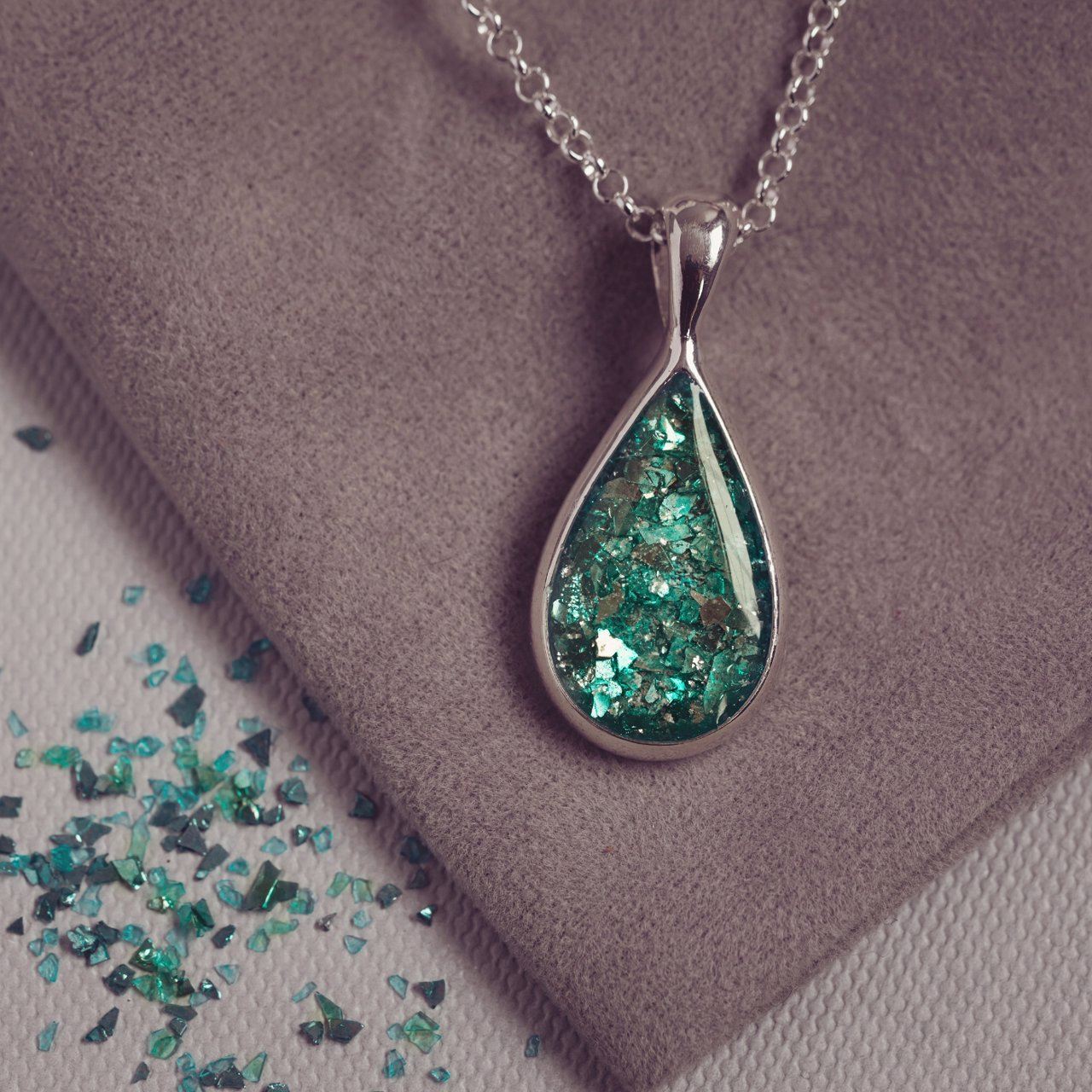 Sterling silver teardrop necklace with a central section filled with memorial ashes and green crushed crystals, displayed on a grey fabric background scattered with green crystal