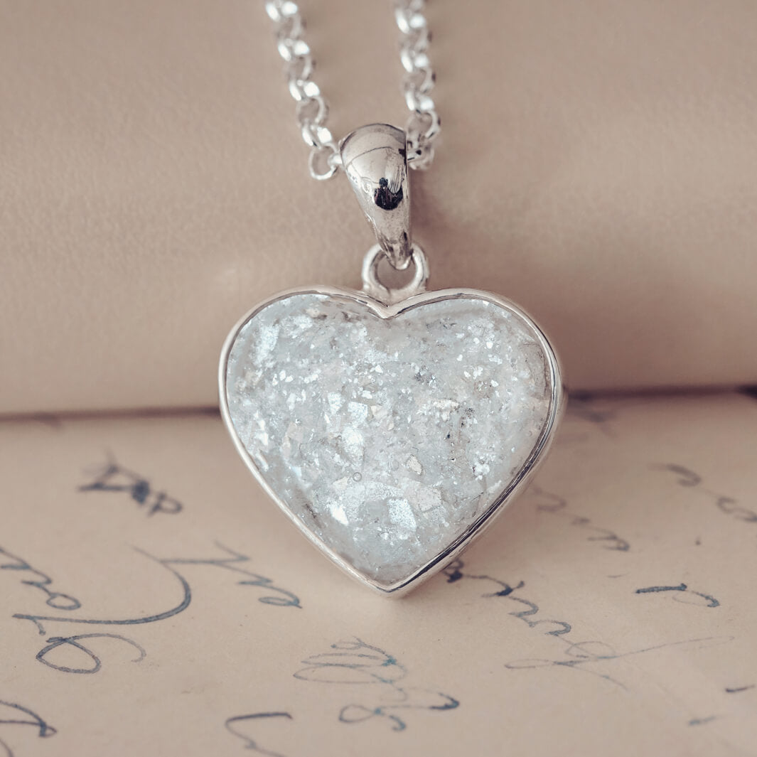 Sterling silver heart necklace with a central section filled with memorial ashes and white & clear crushed crystals, displayed on a beige fabric background.