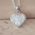 Sterling silver heart necklace with a central section filled with memorial ashes and white & clear crushed crystals, displayed on a beige fabric background.