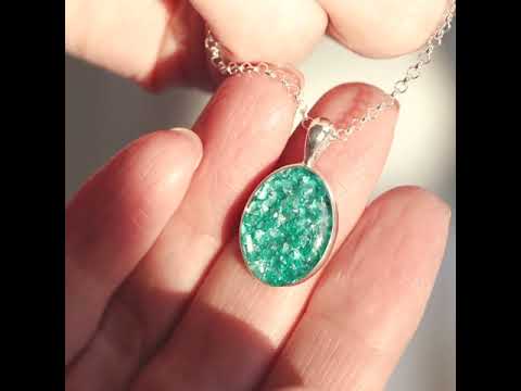 Video of Sterling silver oval necklace with a central section filled with memorial ashes and green crushed crystals, being held and turned by a ladys hands