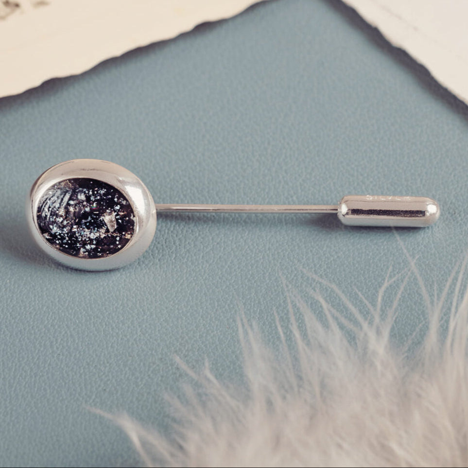 A close up of Memorial Cremation Ashes Tie Pin in black on a blue surface with a grey feather blurred in the corner