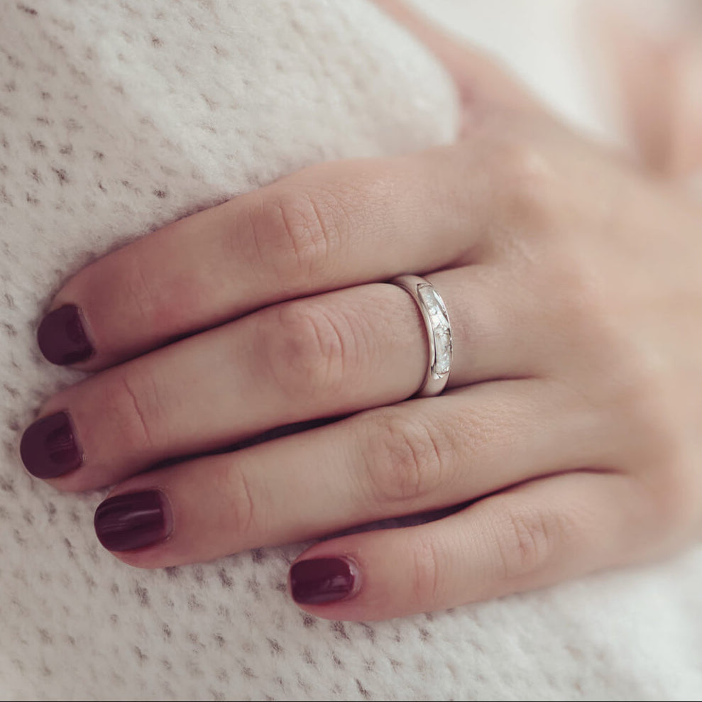 close up of a ladies hand with red painted nails in a white jumper wearing Womens Cremation Ashes Ring Band, the middle of the ring band is filled with clear resin, ashes and white crushed crystals